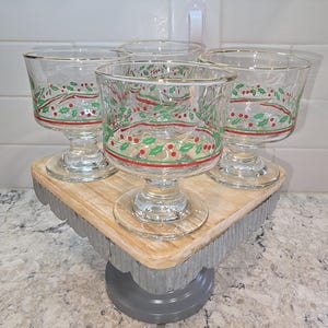 May include: Four clear glass dessert dishes with gold rims and a festive red and green holly berry design. The dishes are displayed on a decorative wooden and metal cake stand, perfect for holiday entertaining.