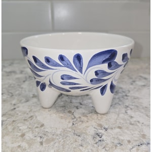 May include: A white ceramic bowl with three short legs, adorned with a blue floral pattern. The bowl's design features stylized blue leaves and swirls, creating a decorative and elegant look. This footed bowl is suitable for serving or display.