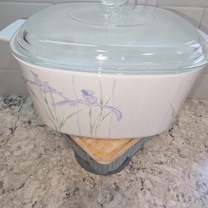 May include: A white casserole dish with a clear glass lid. The dish features a floral design with purple and green accents. The dish is sitting on a wooden trivet.