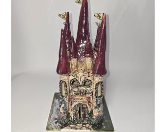 May include: A ceramic castle figurine featuring tall, pointed maroon roofs and a textured beige stone-like base. The structure includes multiple towers with small flags, arched windows, and a central doorway with the word BELIEVE printed above it.