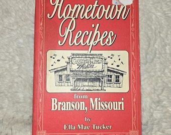 May include: A red cookbook titled Hometown Recipes from Branson, Missouri by Ella Mae Tucker. The cover features a black and white illustration of a Country Music Theatre building with musical notes and a border of kitchen utensil icons.