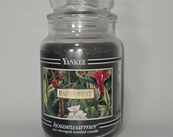 May include: A clear glass jar candle with a lid, containing dark green wax. The label features the text YANKEE, RAINFOREST, and housewarmer our strongest scented candle, along with a colorful illustration of tropical flowers and green foliage.