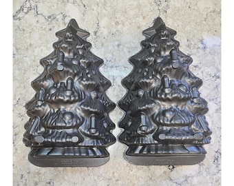 May include: Two dark gray metal cake pans shaped like Christmas trees. Each pan features a molded design of a tree with a star at the top and various circular and cylindrical ornaments embossed into the surface, with a flat base at the bottom.