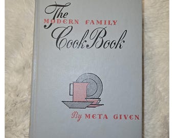 May include: A light gray hardcover book titled The Modern Family Cook Book by Meta Given. The cover features black and pink graphic illustrations of a pitcher, plates, and bowls. The text is printed in a combination of black and pink ink.