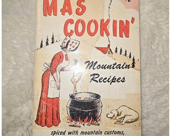 May include: A vintage cookbook titled Ma's Cookin' Mountain Recipes with a cover illustration of a person in a bonnet and apron cooking a chicken in a black pot over a fire, with a dog resting nearby and the text $1.00 at the top right corner.