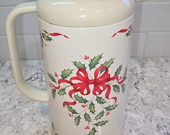 May include: A cream-colored thermal carafe featuring a festive holly and red ribbon pattern. The beverage server has a matching cream-colored lid with a small gold-tone accent on top, a sturdy side handle, and a pouring spout on the upper side.