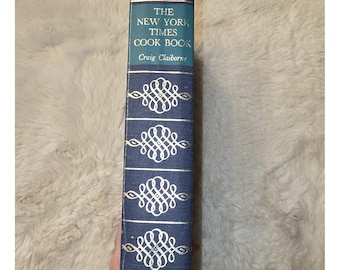 May include: A blue hardcover book titled The New York Times Cook Book by Craig Claiborne, published by Harper & Row. The spine features gold-colored decorative scrollwork patterns and horizontal lines, with the title and author printed in gold.