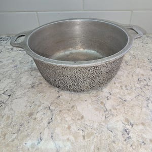 May include: A silver-colored metal cooking pot with two handles. The pot has a textured exterior and a smooth interior. The pot is sitting on a light-colored countertop.