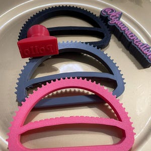 May include: A set of three semi-circular cookie cutters in black, gray, and pink, featuring a serrated outer edge. A small red stamp with the text "BOLLO" is placed on the gray cutter, and a black sign with the text "@lizauracreations" is included.