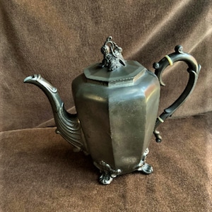 May include: A pewter teapot with a decorative lid and a handle with a wooden accent. The teapot has a hexagonal body and sits on four feet.