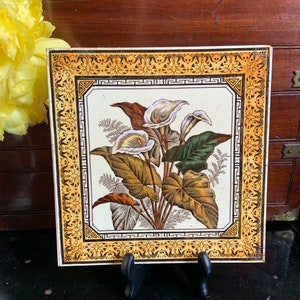 May include: A vintage ceramic tile with a decorative border in yellow and black. The center of the tile features a floral design with white calla lilies and green leaves on a white background.