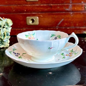 May include: A white teacup and saucer set with floral designs in green, blue, and yellow. The teacup has a curved handle and sits on a matching saucer. The set is placed on a dark surface.