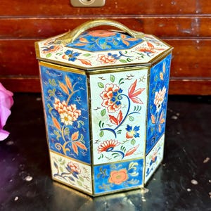 May include: A hexagonal metal tin featuring a decorative floral pattern with alternating panels of deep blue and cream backgrounds. The container includes a matching lid with a gold-toned metal handle and gold-colored trim along the edges.
