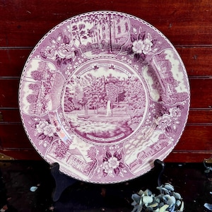 May include: A decorative plate with a purple and white design featuring scenes from Saratoga Springs, NY. The plate has a scalloped edge and floral accents. Text includes "Picturesque Saratoga Springs N.Y." and "Geyser Park Spouting Spring."