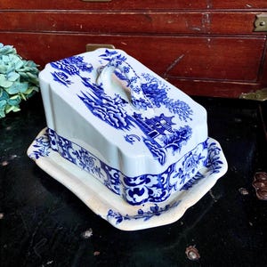 May include: A blue and white ceramic cheese dish with a handle. The dish has a floral design and a raised edge. It is perfect for serving cheese at a party or gathering.