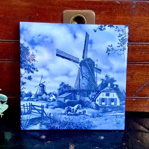 May include: A blue and white ceramic tile with a Dutch windmill scene. The tile features a windmill with a thatched roof, a small house, and a horse and foal in the foreground. The sky is blue with white clouds.