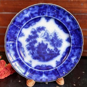May include: A vintage blue and white plate with a detailed landscape scene. The plate features a scalloped edge and intricate patterns. The central design depicts a pagoda, trees, and figures, all in shades of blue against a white background. A decorative piece for display.