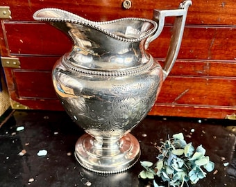 May include: A silver-plated pitcher featuring a beaded rim, a curved handle, and a pedestal base. The body of the pitcher is decorated with an engraved floral pattern, and the spout has a distinct, rounded shape with a beaded edge detail.