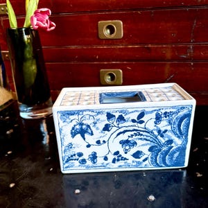 May include: A rectangular ceramic tissue box with a blue and white floral design. The top has a square opening for tissue dispensing. A black glass vase with a pink tulip is to the left.