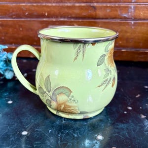 May include: A light green enamel mug with a floral design featuring yellow and brown roses and green leaves. The mug has a silver rim and handle. The mug is a vintage style.