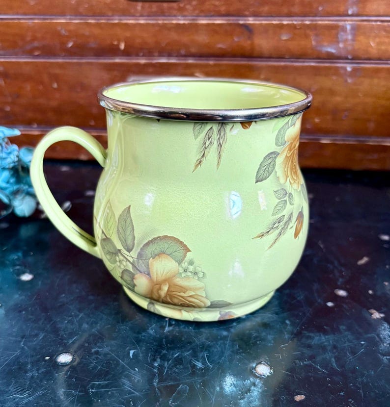 May include: A light green enamel mug with a floral design featuring yellow and brown roses and green leaves. The mug has a silver rim and handle. The mug is a vintage style.