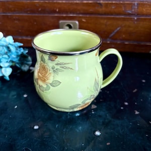 May include: A light green enamel mug with a silver rim and handle. The mug is decorated with yellow and orange rose floral patterns. The mug is sitting on a dark green surface.