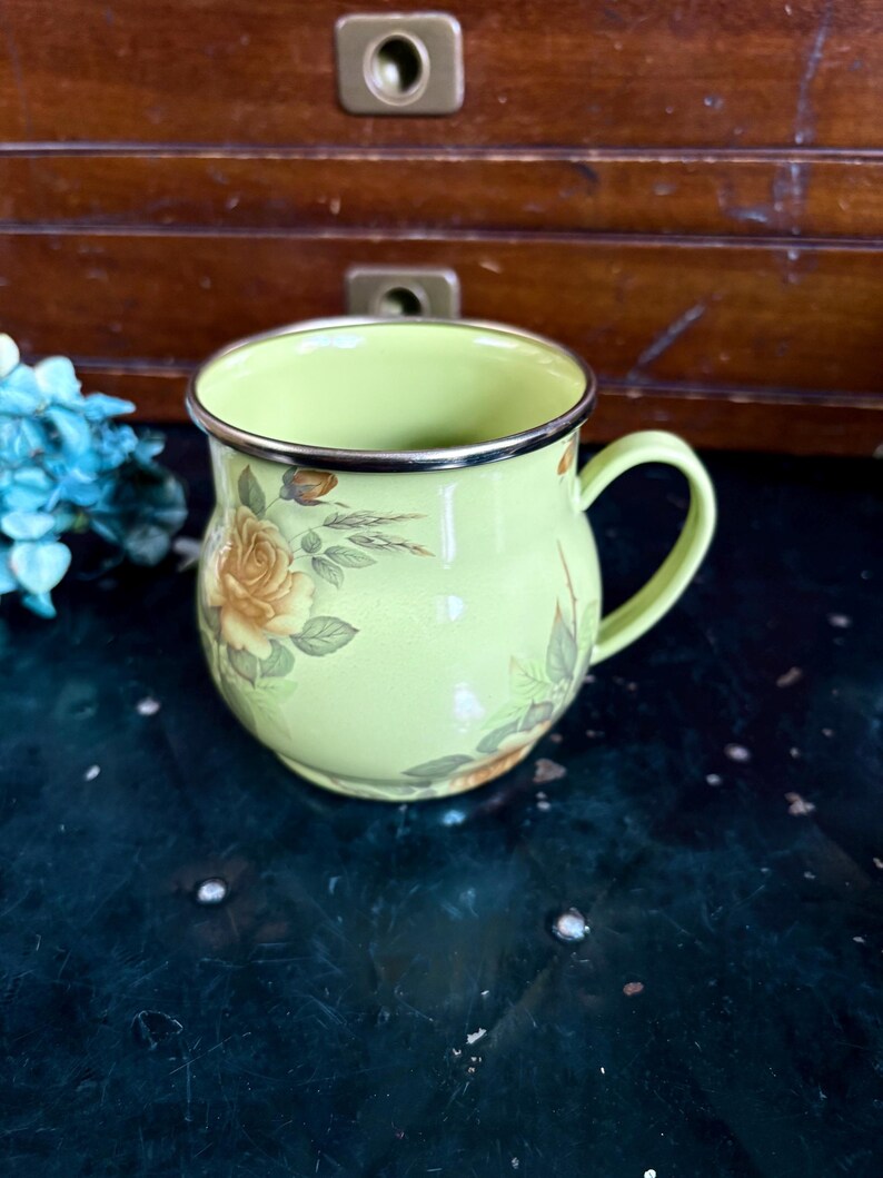 May include: A light green enamel mug with a silver rim and handle. The mug is decorated with yellow and orange rose floral patterns. The mug is sitting on a dark green surface.