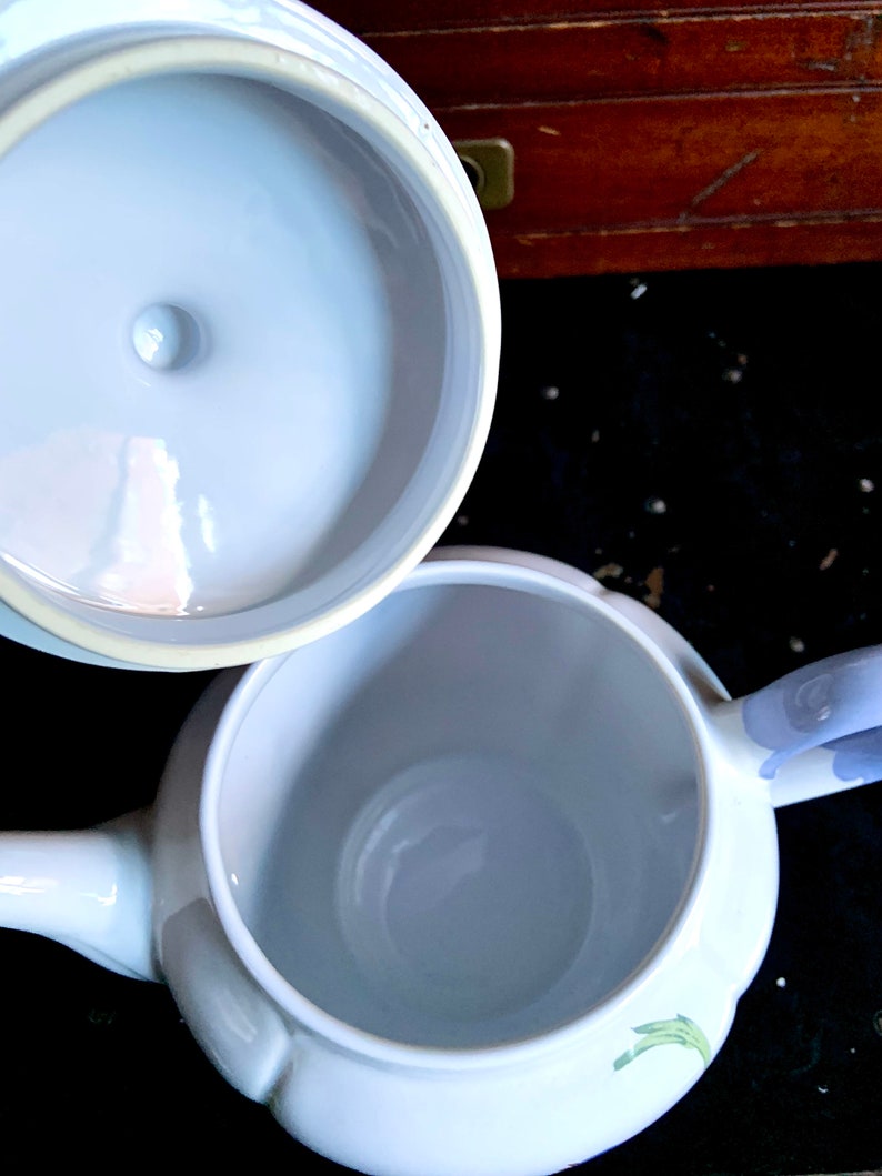 Pu&ograve; includere: Teiera in ceramica bianca con coperchio. La teiera ha un design floreale e un manico. L'interno della teiera &egrave; visibile.