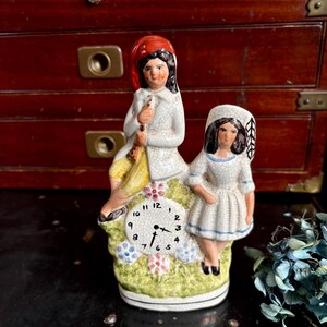 May include: A ceramic figurine featuring two figures atop a clock face. One figure wears a red hat and white coat, holding a flute. The other figure is in a white dress with blue trim. The clock has black numbers.