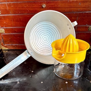 May include: A vintage kitchen scene featuring a metal strainer with a long handle and a yellow citrus juicer. The juicer has a clear glass base and a bright yellow top. The strainer is silver and has many small holes.