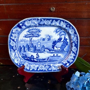 May include: An oval ceramic platter featuring a blue and white transferware pattern. The central scene depicts a landscape with a river, a bridge, a small house, and figures in boats. The border is decorated with a repeating floral motif.