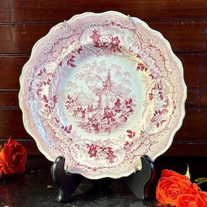 May include: A large, antique, scalloped-edge plate with a red and white floral and landscape design. The plate is supported by a black stand, and the background features a dark wooden surface. The plate's design includes floral patterns and a scenic landscape.