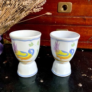 May include: Two white ceramic egg cups with a blue, yellow, and purple bird design. The egg cups have a flared base and a blue trim. A bundle of dried lavender is in the background.