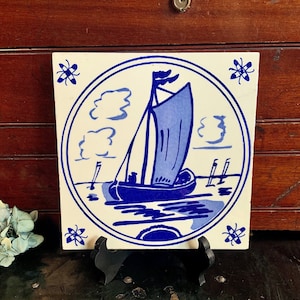 May include: A blue and white ceramic tile with a hand-painted design of a sailboat on a body of water. The tile is surrounded by a blue border and has a white background. The tile is in a circular frame with a blue border.
