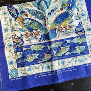 May include: A blue and white Irish linen textile featuring a parrot design. The textile has a blue border and includes text that reads "Inspired by William De Morgan for the Fitz William Museum". The design includes fish and floral elements.