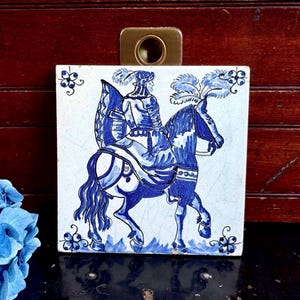 May include: Square ceramic tile with a blue and white illustration of a person on horseback. The tile has a brass clip on the top. The artwork features a detailed depiction of the rider and horse, with floral accents.