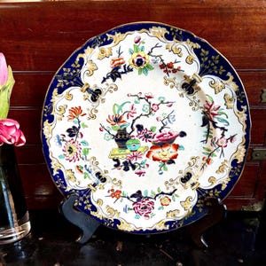 May include: Antique decorative plate with a white center featuring a colorful floral design. The plate has a dark blue border with gold accents and a scalloped edge. The plate is displayed on a black stand.