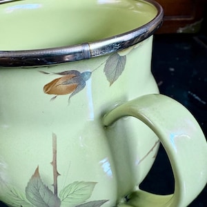 May include: A vintage enamelware pitcher in a pale green hue, adorned with floral and leaf motifs in muted tones. The pitcher features a silver rim and a curved handle, showcasing a classic design. This piece is ideal for kitchen decor.