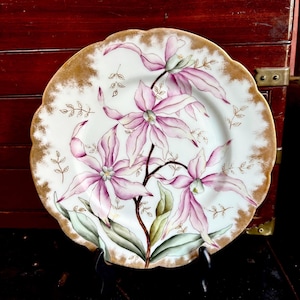 May include: A white porcelain plate with a scalloped gold rim. The plate features a hand-painted floral design with pink and purple orchids and green leaves. The plate is displayed on a black stand.