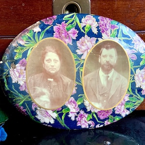 May include: Oval-shaped decorative plate with a blue background and a floral pattern. Two oval-shaped portraits of a man and a woman are framed in gold and set against the floral background.
