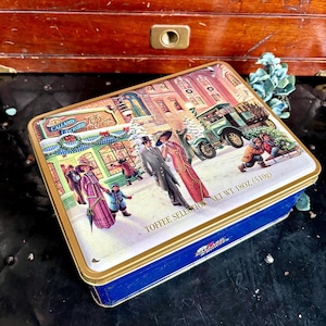 May include: A rectangular tin with a winter scene illustration. The lid features a vintage Christmas scene with people, a car, and a shop. The tin is blue with a gold trim and the text "Toffee Selection Net Wt 18oz (510g)".