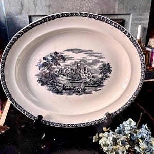 May include: An oval, off-white serving platter with a black and white landscape scene. The rim features a decorative black border. The landscape depicts a river, boats, trees, and buildings. A classic piece for serving food.