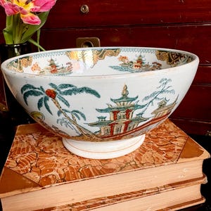 May include: A white porcelain bowl with a wide rim and a pedestal base. The bowl is decorated with a colorful Asian-inspired landscape scene featuring pagodas, palm trees, and boats. The bowl is resting on a stack of old books.