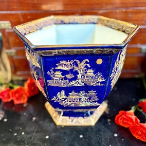 May include: A blue and gold ceramic planter with an octagonal shape. The planter features a gold Asian-inspired landscape design on a dark blue background. The rim and base are also trimmed in gold. Red roses are in the foreground.