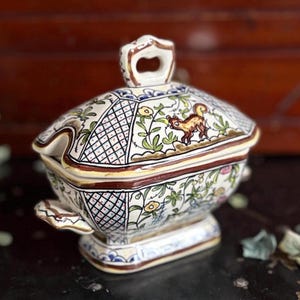 May include: A decorative ceramic serving dish with a lid. The dish is white with a hand-painted floral and squirrel design in brown, green, yellow, and blue. The lid has a handle and the dish has two side handles.