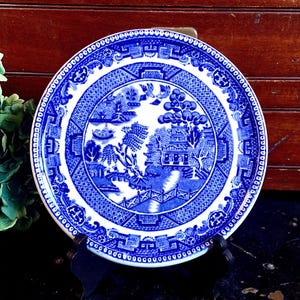 May include: A blue and white porcelain plate with a willow pattern design. The plate features a traditional Chinese landscape scene with a bridge, trees, and a pagoda.