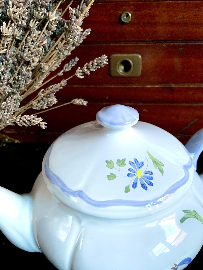 Pu&ograve; includere: Teiera bianca con un motivo floreale blu e verde. La teiera ha un coperchio e un beccuccio. La teiera &egrave; appoggiata su una superficie nera.