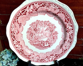 May include: A large, white ceramic plate with a pink floral design. The plate features a central scene of a bridge and trees, with figures in the foreground. The plate has a scalloped edge.
