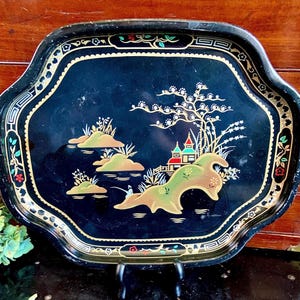May include: A black metal serving tray with a gold rim and a painted scene of a Japanese garden with a pagoda, trees, and water. The tray has a scalloped edge.