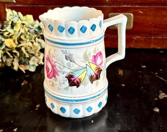 May include: A vintage white ceramic pitcher with a handle, adorned with pink roses and blue diamond accents. The pitcher has a scalloped rim and a decorative blue band. The overall design is ornate and floral.
